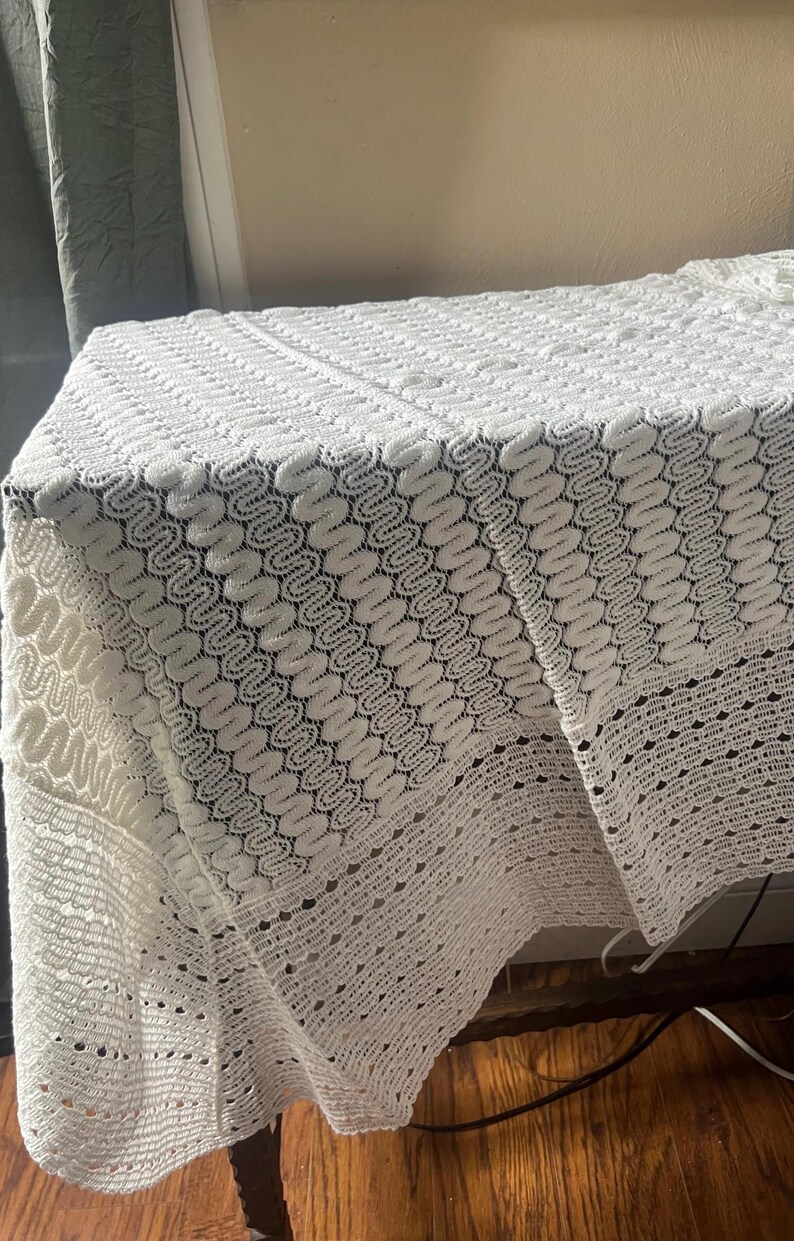 May include: A white, intricately patterned lace tablecloth. The tablecloth is draped over a table, showcasing a detailed, openwork design. The lace features a repeating, wave-like pattern, adding texture and visual interest to the textile.
