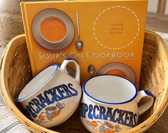 Peut inclure: Un panier en bois tressé contenant un livre de cuisine jaune intitulé Soup's On Cookbook et deux tasses en céramique blanche à bord bleu. Les tasses portent l'inscription Soup & Crackers en bleu, avec des illustrations de légumes et de crackers.