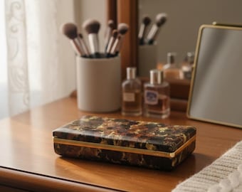 Peut inclure: Une boîte décorative rectangulaire avec un motif floral marron foncé et doré, bordée d'or. La boîte est posée sur une coiffeuse en bois, avec des pinceaux de maquillage, des flacons de parfum et un petit miroir.