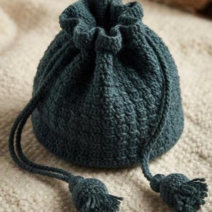 Puede incluir: Una bolsa de ganchillo de color verde azulado oscuro con cordón y un patrón de puntos texturizados. La bolsa tiene una abertura superior fruncida asegurada por dos cordones de hilo a juego, cada uno con una pequeña borla redonda.