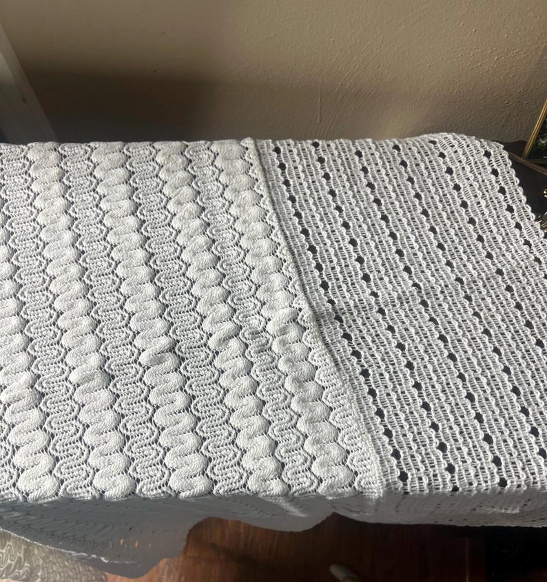 May include: A white, textured, knitted blanket with a detailed pattern. The blanket is divided into two sections, one with a wavy design and the other with a dotted pattern. The blanket is made of a soft material and is suitable for home decor.