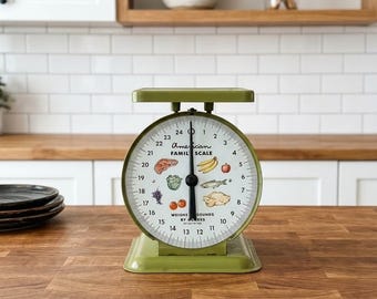Peut inclure: Une balance de cuisine analogique vert olive avec un cadran rond portant l'inscription American Family Scale et des illustrations de divers aliments comme de la viande, des bananes, une pomme, du poisson, de la volaille, des tomates, du raisin et de la laitue, avec une aiguille noire.