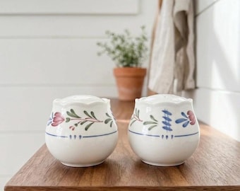 Peut inclure: Une paire de salière et poivrière en céramique blanche de forme arrondie avec des bords supérieurs festonnés. Chaque pièce présente un motif floral peint à la main dans des tons rouge, vert et bleu, avec une fine ligne bleue horizontale.