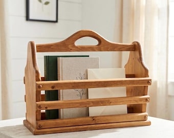 Peut inclure: Un porte-revues en bois de ton moyen doté d'une poignée centrale découpée, de deux lattes horizontales sur le devant et d'une base solide. Il contient un livre vert foncé, un livre beige clair avec une illustration de feuille et une enveloppe crème.