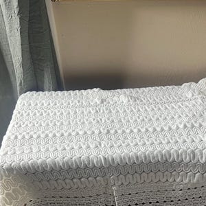 May include: A white, textured fabric with a repeating wave pattern. The fabric is draped over a rectangular form, with the edges of the fabric showing a scalloped design. The background includes a gray curtain and a neutral-colored wall.