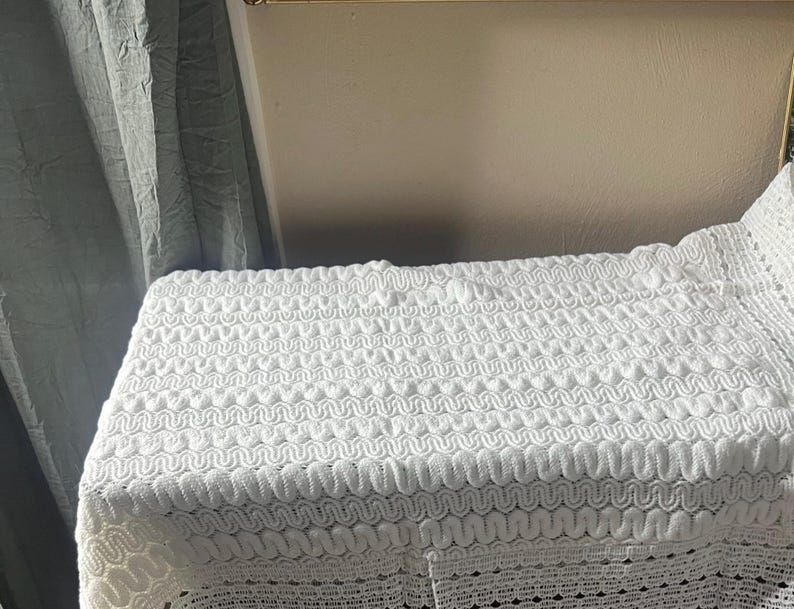 May include: A white, textured fabric with a repeating wave pattern. The fabric is draped over a rectangular form, with the edges of the fabric showing a scalloped design. The background includes a gray curtain and a neutral-colored wall.