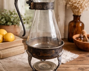 Peut inclure: Une carafe en verre transparent avec une bande et une poignée en métal décoratif, placée dans un support en métal vintage. La carafe est sur une surface en bois, avec des citrons et des livres en arrière-plan. La carafe est de style vintage.