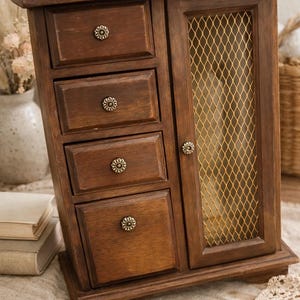 May include: A wooden jewelry armoire with a dark brown finish. It features four small drawers with decorative knobs and a door with a gold mesh panel. The armoire is designed to store jewelry and accessories.