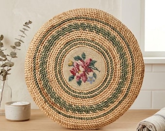 Peut inclure: Une décoration murale ronde en jonc de mer tressé, composée de cercles concentriques de fibres couleur beige naturel et vert sauge. Le centre présente un insert circulaire en tissu beige brodé d'un motif floral rose, rouge et vert.