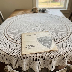 May include: A cream-colored, 70-inch crocheted cloth tablecloth with a detailed floral design. The tablecloth is laid on a wooden table, with a vintage pattern for the cloth visible. The pattern is labeled "Design 7172".