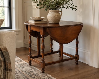 Peut inclure: Une table à abattants en bois brun foncé avec des pieds tournés et un plateau rectangulaire. La table comporte deux battants latéraux articulés qui peuvent être abaissés. Un vase en céramique beige avec du feuillage et un bol sur un livre.