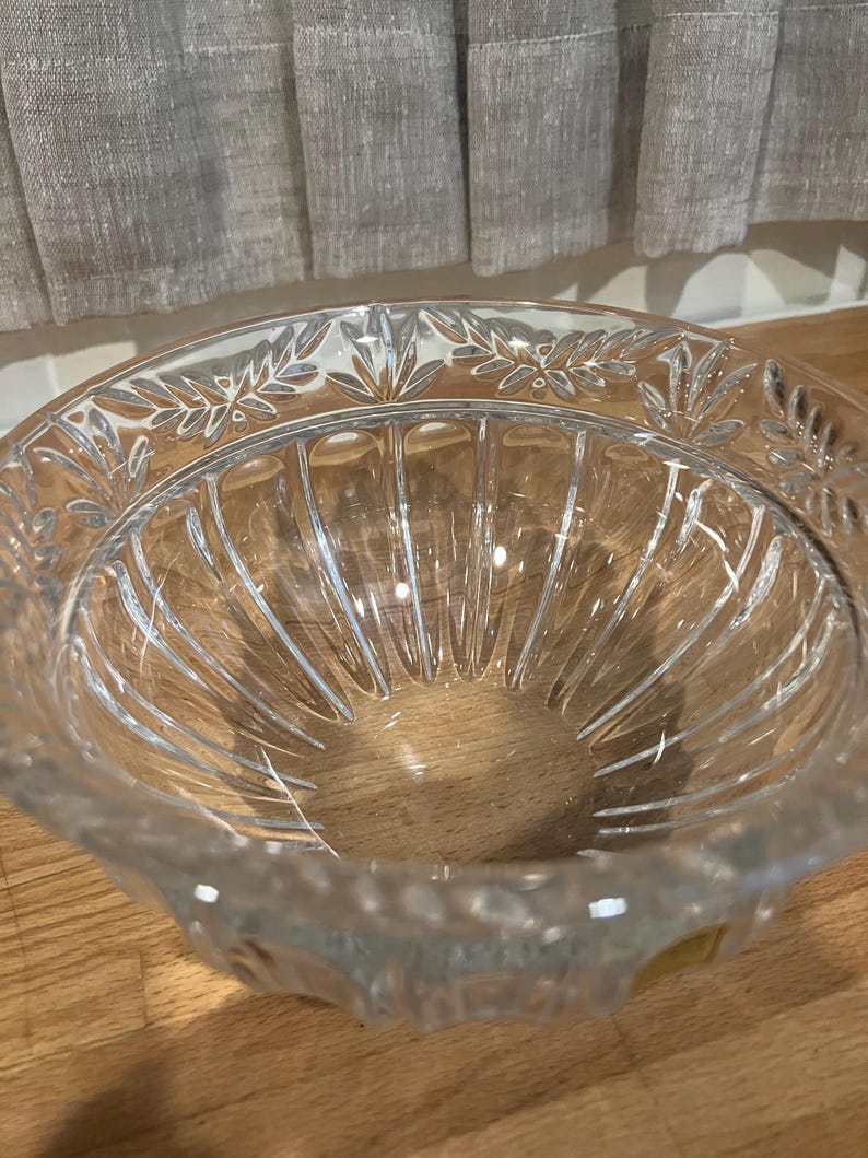 May include: A clear glass bowl with a decorative leaf pattern around the rim. The bowl has vertical ridges on the inside and sits on a wooden surface. The glass is transparent, allowing light to pass through.