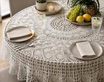 May include: A round, cream-colored crocheted tablecloth covers a dining table set for a meal. The table is set with plates, silverware, and glasses. A centerpiece of fruit and a small potted plant add to the decor.