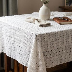 May include: A white, intricately crocheted tablecloth covers a wooden table. A beige vase with greenery, a folded napkin, and a leather-bound notebook with a pen rest on the table. The tablecloth has a detailed, textured pattern.