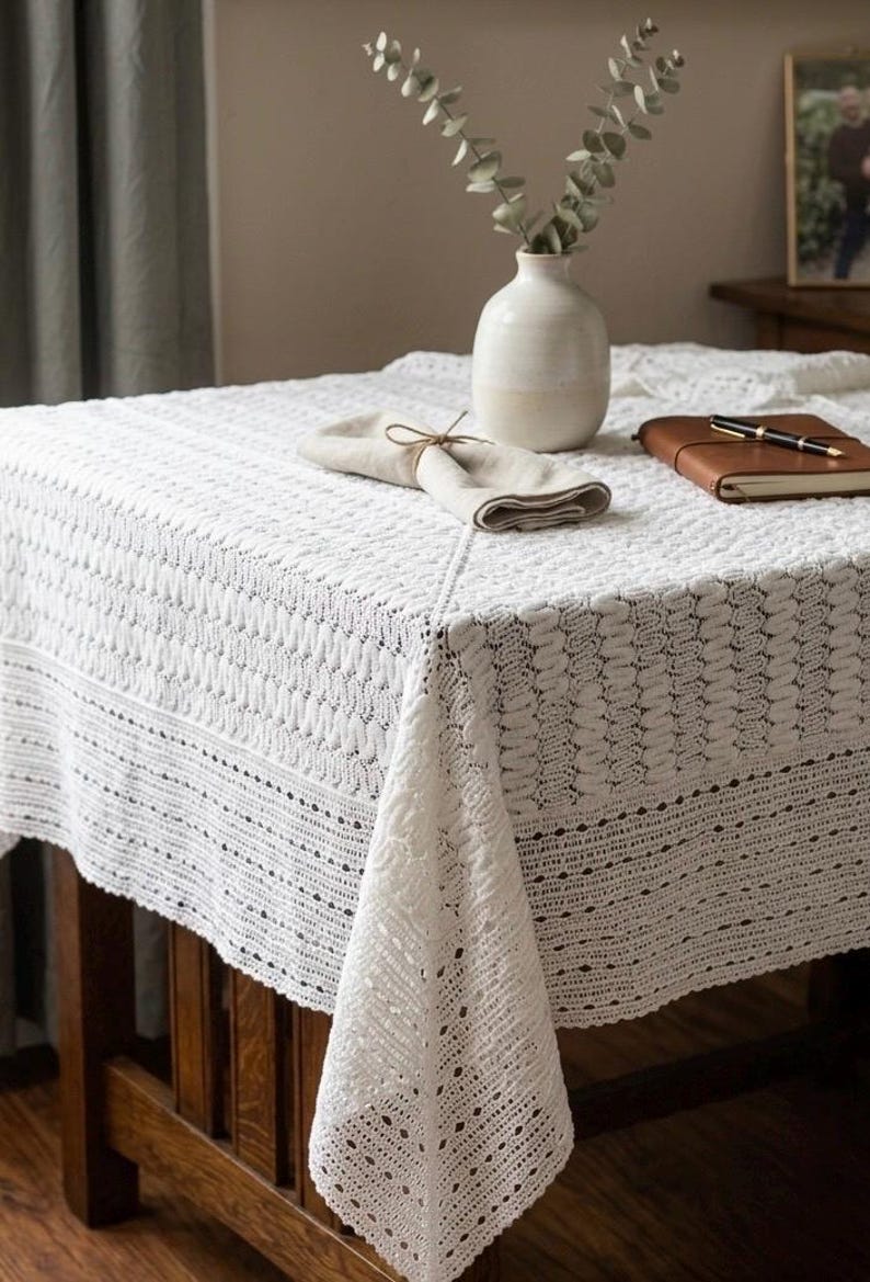 May include: A white, intricately crocheted tablecloth covers a wooden table. A beige vase with greenery, a folded napkin, and a leather-bound notebook with a pen rest on the table. The tablecloth has a detailed, textured pattern.