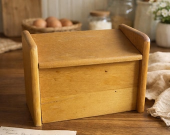 Peut inclure: Un coffre à pain en bois brun clair avec un couvercle incliné et des supports latéraux incurvés. Le coffre est en bois et a une finition naturelle. Le coffre à pain est posé sur une surface en bois.
