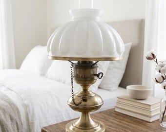 Peut inclure: Une lampe de style à huile en laiton avec un abat-jour en verre blanc cannelé et une cheminée en verre transparent. La lampe possède une base métallique avec un brûleur central, une petite chaînette et une clé de réglage latérale.