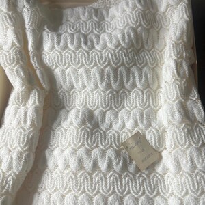 May include: A white, knitted garment with a repeating wave pattern. The garment is folded inside a cardboard box. A small tag is attached to the garment. The knitted material has a textured, three-dimensional appearance.