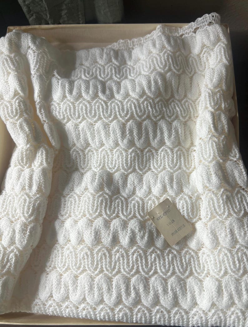 May include: A white, knitted garment with a repeating wave pattern. The garment is folded inside a cardboard box. A small tag is attached to the garment. The knitted material has a textured, three-dimensional appearance.