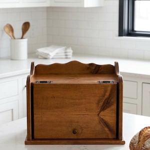 May include: A medium-brown wooden bread box with a hinged front door, a small circular knob, and a scalloped top edge. The box features visible wood grain patterns and two metal hinges on the top, providing a storage container for baked goods.