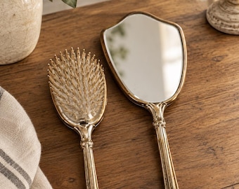 Peut inclure: Une brosse à cheveux et un miroir à main assorti de style vintage avec des manches métalliques dorés ornés. La brosse présente une disposition dense de poils, tandis que le miroir a une surface réfléchissante et une forme en écusson.