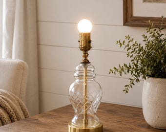 Peut inclure: Une lampe de table avec une base en verre transparent ornée d'un motif en losanges, un col en métal finition laiton et un socle circulaire en laiton. La lampe présente une douille apparente surmontée d'une ampoule blanche allumée.