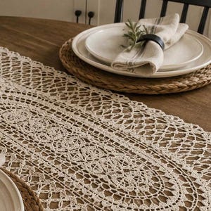 May include: An off-white, oval-shaped crochet table runner featuring an intricate, openwork lace pattern with scalloped edges. The runner is laid flat on a wooden table surface, extending across the center between two woven placemats.