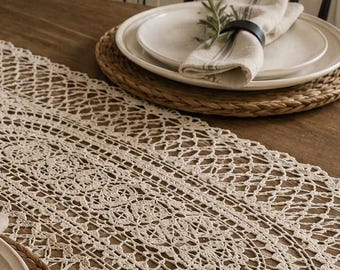 May include: An off-white, oval-shaped crochet table runner featuring an intricate, openwork lace pattern with scalloped edges. The runner is laid flat on a wooden table surface, extending across the center between two woven placemats.