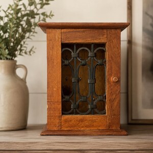 May include: A rectangular wooden cabinet with a natural brown finish featuring a central door. The door is inset with a black metal grate composed of vertical oval shapes and circular connectors. A small wooden knob is attached to the right side.