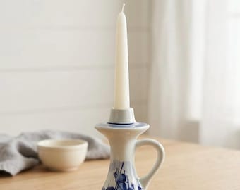 Peut inclure: Un bougeoir en céramique blanche orné d'un motif de moulin à vent peint en bleu, doté d'une anse, d'une base évasée et d'un col étroit. Une bougie fine de couleur blanc cassé est insérée dans l'ouverture supérieure du bougeoir.