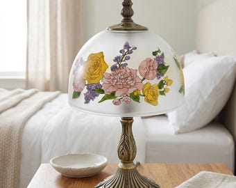 Peut inclure: Une lampe de table avec un abat-jour en verre dépoli en forme de dôme orné d'un imprimé floral coloré de roses jaunes, de pivoines roses et de touches violettes. La base en métal couleur laiton antique présente un design cannelé.