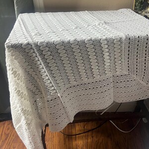May include: A white, crocheted tablecloth with a detailed, openwork pattern. The square tablecloth is draped over a table, showcasing the intricate design and texture of the handmade piece. The edges feature a scalloped border.