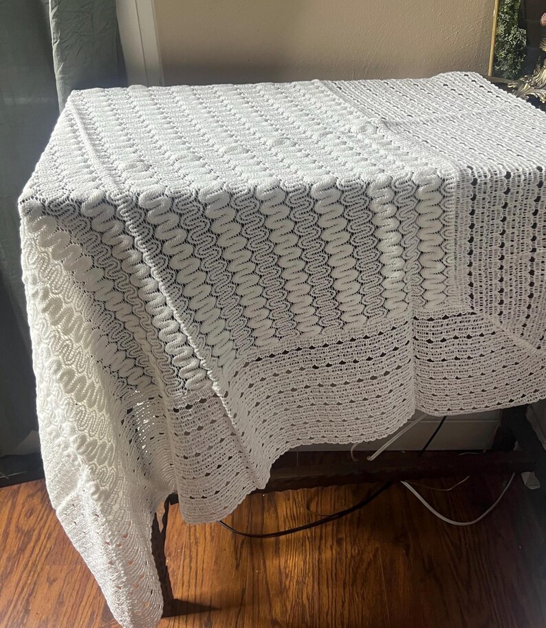 May include: A white, crocheted tablecloth with a detailed, openwork pattern. The square tablecloth is draped over a table, showcasing the intricate design and texture of the handmade piece. The edges feature a scalloped border.