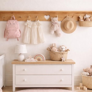 May include: A light-filled nursery scene with a wooden shelf holding a pink backpack, a white dress, a teddy bear, and a straw hat. A white dresser with two drawers is below, topped with a lamp, books, and a rainbow. Wicker baskets and a crib complete the decor.