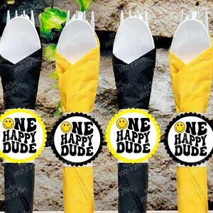 May include: Four yellow and black napkin rings with the text "One Happy Dude" and a smiley face. The rings are placed around white forks and napkins.