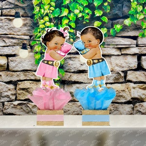 May include: Two adorable baby figures, one in a pink dress and the other in a blue outfit, are holding boxing gloves. They are standing on top of decorative boxes with tissue paper, one pink and one blue.