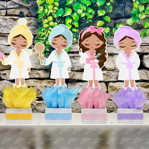 May include: Four spa-themed centerpieces featuring cartoon figures wearing bathrobes and holding spa items. Each centerpiece has a tissue paper base in a different color: yellow, blue, pink, and purple.