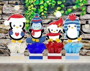 May include: Four penguin-themed centerpieces with various winter hats and scarves. Each penguin is perched atop a box with tissue paper in white, blue, and red. The background features a stone wall and greenery.