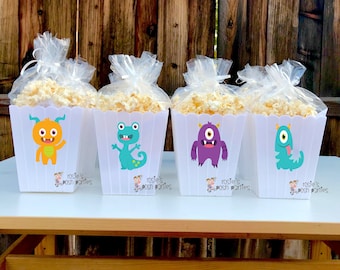May include: Four white popcorn containers with monster designs. Each container has a different monster with a unique color scheme. The containers are filled with popcorn and have a clear plastic bag tied around the top. The containers have the text "Rosie's posh parties" printed on the bottom.