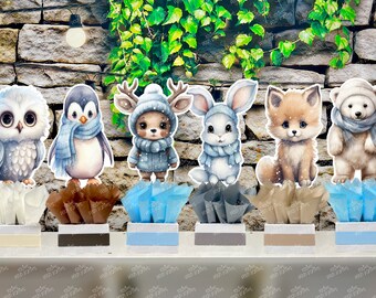 May include: Six adorable animal centerpieces for a winter-themed party. Each centerpiece features a different animal, including an owl, penguin, deer, bunny, fox, and polar bear. The animals are all wearing winter scarves and hats, and they are sitting on top of small, decorative bases.