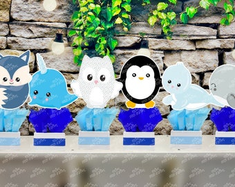 May include: A collection of six arctic animal-themed centerpieces. Each centerpiece features a different animal: a fox, narwhal, owl, penguin, seal, and whale. The animals are set on blue tissue paper and white and blue bases, perfect for a winter-themed party.