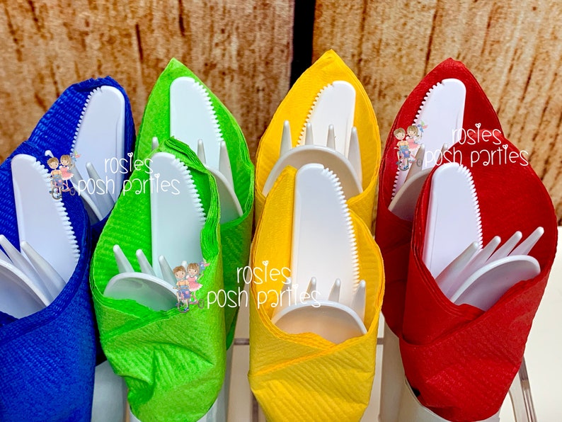 Op de afbeelding: Vier sets witte plastic bestek verpakt in kleurrijke servetten. De servetten zijn blauw, groen, geel en rood. Het bestek bevat een mes, vork en lepel.