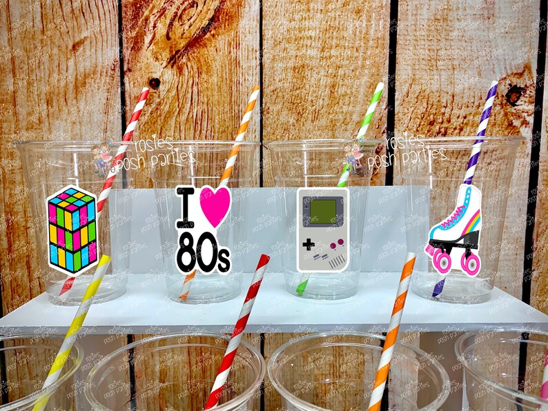 May include: Six clear plastic cups with colorful straws and 80s themed stickers. The stickers feature a Rubik's Cube, a heart with "I Love 80s" text, a retro video game console, and roller skates.