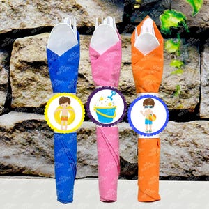 Op de afbeelding: Drie servetringen in blauw, roze en oranje met een gegolfde rand. Elke ring heeft een ander ontwerp: een jongen in een zwemvest, een emmer en een schep en een jongen met een strandbal.