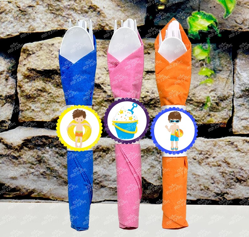 Op de afbeelding: Drie servetringen in blauw, roze en oranje met een gegolfde rand. Elke ring heeft een ander ontwerp: een jongen in een zwemvest, een emmer en een schep en een jongen met een strandbal.
