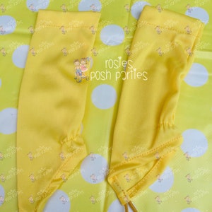 May include: Two yellow arm warmers with elastic bands at the bottom. The arm warmers are made of a smooth fabric and have a small embroidered design of two children on a bicycle. The background is a yellow polka dot pattern.