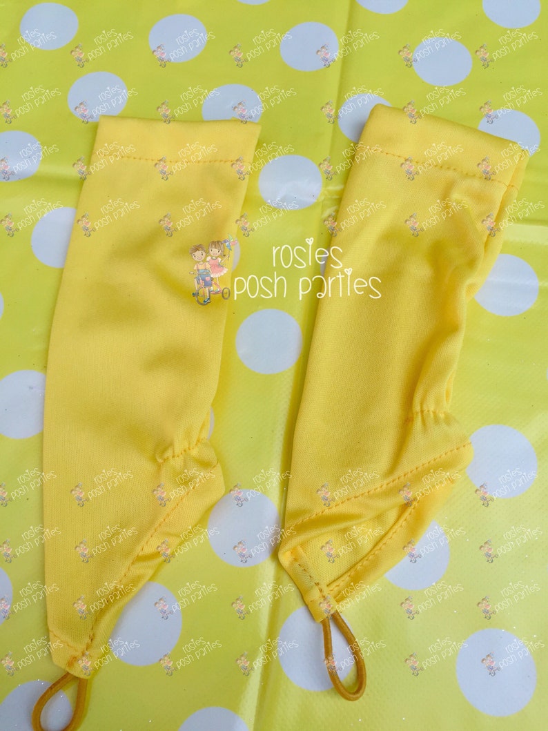 May include: Two yellow arm warmers with elastic bands at the bottom. The arm warmers are made of a smooth fabric and have a small embroidered design of two children on a bicycle. The background is a yellow polka dot pattern.