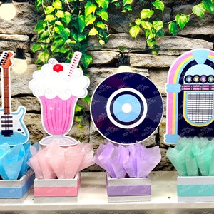 May include: Four centerpieces featuring a blue guitar, a pink milkshake, a black vinyl record, and a blue and pink jukebox. Each centerpiece is placed in a white box with a colored tissue paper base.