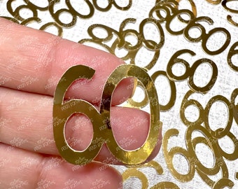 May include: Gold metallic confetti in the shape of the number 60 scattered on a white surface. The number 60 is repeated multiple times. A hand is holding a gold number 60. The confetti is for a birthday or anniversary celebration.