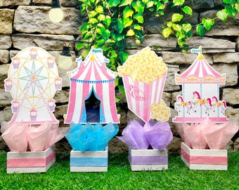 May include: Four circus-themed centerpieces. The designs include a Ferris wheel, a circus tent, a popcorn box with the text "Pop Corn", and a carousel. Each centerpiece sits in a white box with pink, blue, or purple accents and tissue paper.